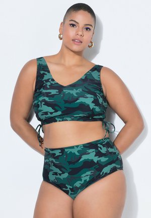 Vrouw met kort haar, gekleed in een groene en zwarte camouflerende bikinibroek met hoge taille, staand tegen een witte achtergrond met haar handen achter haar rug.