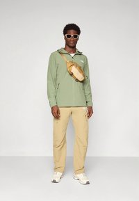 Lysegrønn jakke, beige bukser og hvite joggesko. Jakken har glidelås og lommer. En brun crossbody-veske bæres på tvers av brystet.