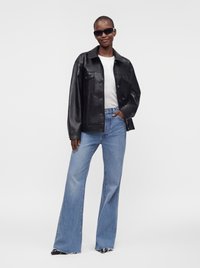 Personne portant une veste en cuir noire, une chemise blanche, un jean bleu à jambes larges, des chaussures noires pointues et des lunettes de soleil foncées, debout sur un fond uni.