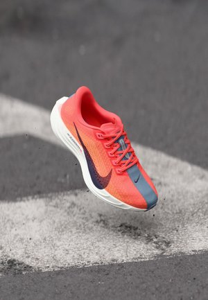 Heller orange und blauer Nike-Laufschuh in der Luft über Asphalt mit weißen Straßenmarkierungen, der Details von Schnürsenkeln und Sohle zeigt.