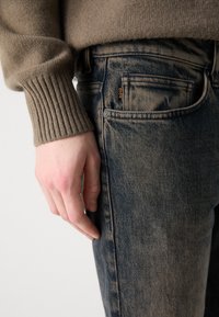 Suéter de punto marrón con puños acanelados, combinado con jeans de mezclilla oscuros y descoloridos que cuentan con una etiqueta de marca naranja visible en el bolsillo.