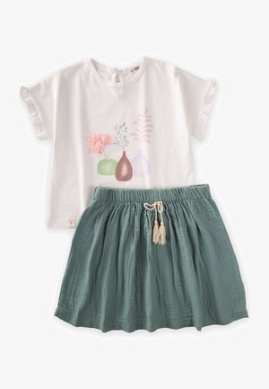 White short-sleeve top with floral print and ruffled edges paired with a green cotton skirt featuring an elastic waistband and tassel detail.