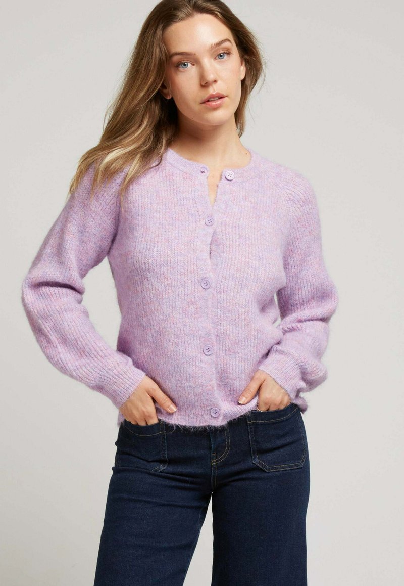 Cardigan lilas avec une texture douce et pelucheuse, présentant un col rond, des manches longues et six boutons assortis sur le devant. Stylisé avec un jean foncé.