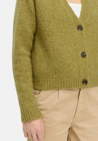 Olijfgroen, gebreide cardigan met een V-hals, ribbelzoom en lange mouwen. Heeft twee donkerbruine knopen aan de voorkant.