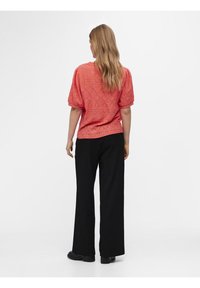 Koraalroze oogjesblouse met pofmouwen, gecombineerd met zwarte wijde broekspijpen. De blouse heeft een gestructureerde, gedessineerde stof.