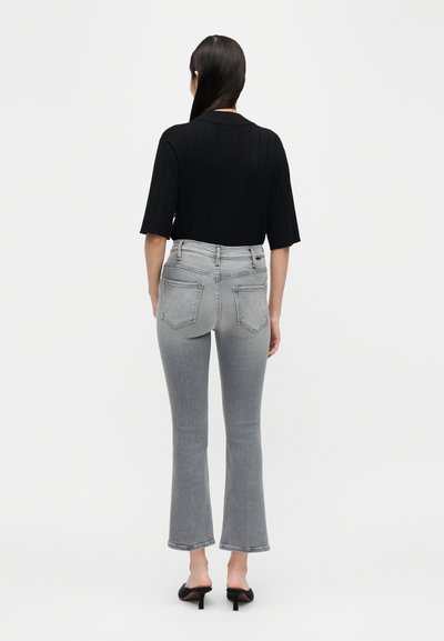 Γυναίκα με μακριά σκούρα μαλλιά, φοράει μαύρο κοντομάνικο τοπ, γκρι τζιν καμπάνα με ψηλή μέση και μαύρα σανδάλια με τακούνι, στέκεται με πλάτη.