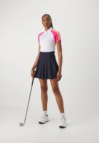 Fehér és rózsaszín golfpóló rövid ujjakkal; hozzá egy pliszírozott tengerészkék szoknya; golfütőt tart, fehér sportcipőt visel.