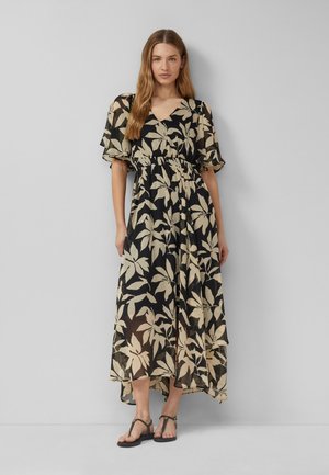 Vrouw staand, een zwarte maxi-jurk met beige bloemenprint, korte vlindermouwen en zwarte platte sandalen dragend.