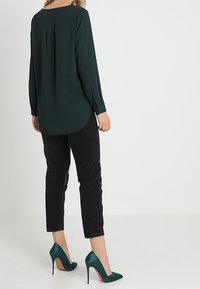 Blusa lunga a maniche verdi scuri con scollatura rotonda e piega sul retro, abbinata a jeans neri e scarpe con tacco teal.