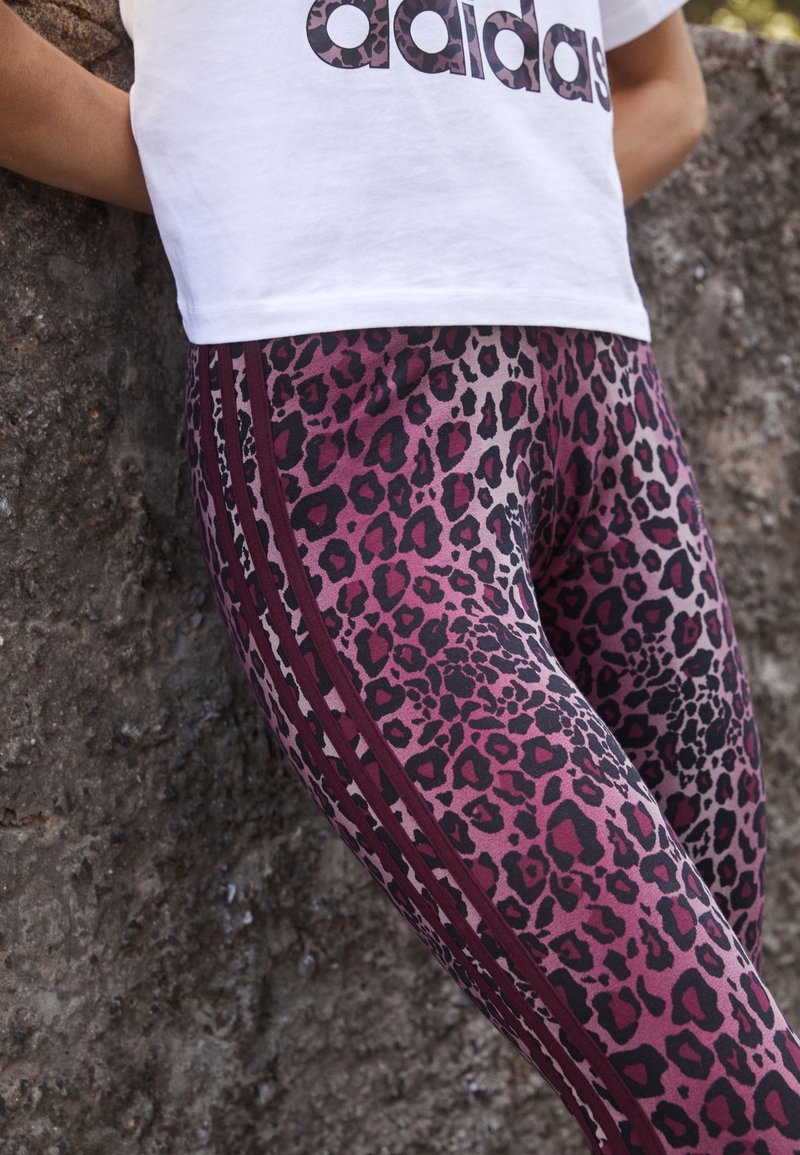 Person wearing purple and black leopard print leggings with maroon side stripes and a white Adidas logo t-shirt, leaning against a stone wall.