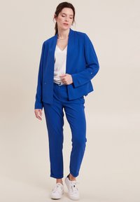 Blazer blu su misura con top bianco a scollo a V e pantaloni blu. L'outfit presenta una texture liscia ed è abbinato a sneaker bianche.