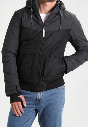 Mand iført sort og mørkegrå hættetrøje med lynlås foran og blå jeans, hånd i lommen.