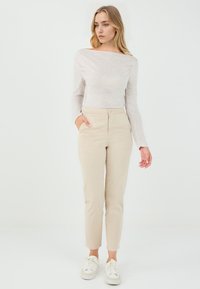 Lichtbeige broek met een aansluitende pasvorm, hoge taille, voorzakken en een knoopsluiting. Gepaard met een lichtgrijs shirt met lange mouwen.
