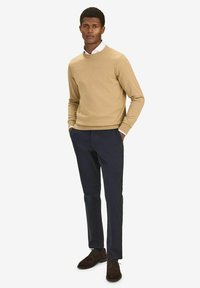 Beige teksturoitu neule, tummansiniset slim-fit-housut ja tummanruskeat mokkakengät. Malli seisoo kädet taskuissa yksinkertaista taustaa vasten.