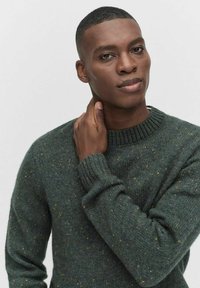 Donkergroen gebreid sweater met geribbelde manchetten en halslijn, voorzien van een gespikkelde structuur met kleine gele en bruine vlekken.