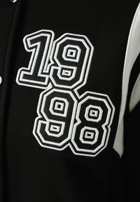 Černý dres s bílými vyšívanými čísly "1998" s výrazným obrysem. Měkká tkanina s kontrastními panelem. Zapínání na patentky.