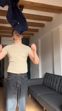 Muž v interiéri hádže tmavú bundu nahor, stojaci pri sivej pohovke s drevenými trámami na strope nad hlavou.