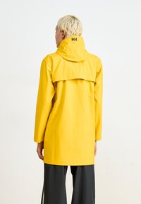 Chaqueta de lluvia amarilla impermeable con capucha, que cuenta con un yugo trasero y detalles de botones a presión. La tela parece suave y duradera.