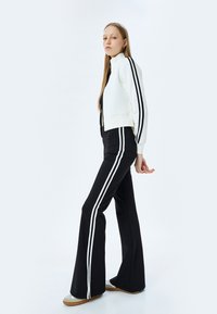 Witte zip-up jas met zwarte strepen, gecombineerd met zwarte flared broek voorzien van witte zijstrepen. De stof lijkt soepel en rekbaar.