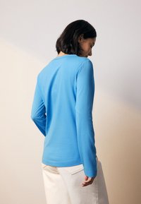 Modra srajca z dolgimi rokavi, z okrolim izrezom, gladke teksture in oprijetega kroja, kombinirana s svetlimi hlačami.