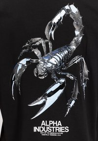 Γραφικό μεταλλικού ασημί σκορπιού με ανυψωμένα νύχια σε μαύρο ύφασμα πάνω από λευκό κείμενο "Alpha Industries" και λεπτομέρειες τοποθεσίας.