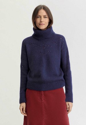 Pull en tricot bleu marine avec col roulé, ourlet côtelé et motifs discrets en œillets. Assorti à une jupe en velours côtelé rouge.