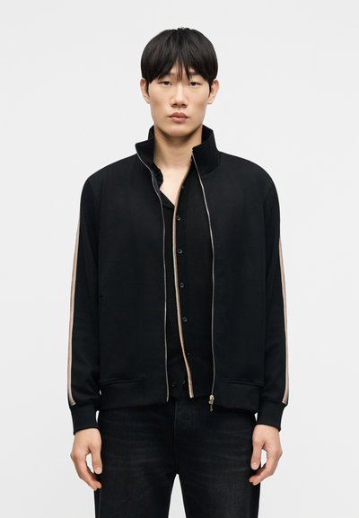 Μαύρο τζάκετ με φερμουάρ και ψηλό γιακά, με πολύχρωμες ριγέ λεπτομέρειες κατά μήκος των μανικιών. Φέρει μαύρο πουκάμισο με κουμπιά από κάτω.