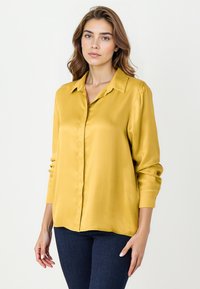 Żółta satynowa koszula na guziki z kołnierzykiem, długimi rękawami i prostym dołem, wystawiona na ciemnych dżinsowych spodniach. Gładka tekstura.