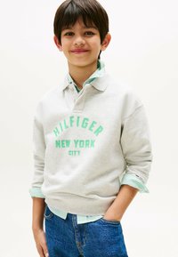 Felpa grigia con stampa verde "HILFIGER NEW YORK CITY", indossata sopra una camicia a righe verde chiaro, abbinata a jeans in denim blu.