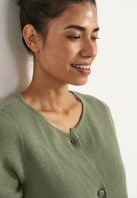 Grön stickad cardigan med rund halsringning, framknappar och textilstruktur. Närbild på en modell med ett blyertssmil.
