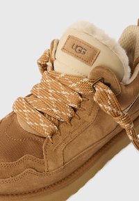 Sneaker din suede tan și plasă, cu suprafață texturată, șireturi late și căptușeală pufoasă. Include o etichetă cu logo-ul UGG pe gulerul căptușit.
