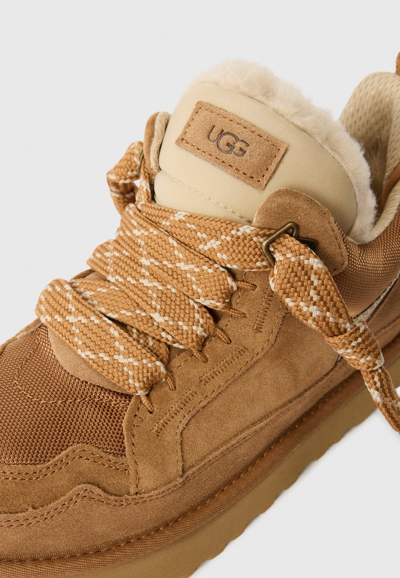 Beige Wildleder- und Mesh-Sneaker mit strukturiertem Obermaterial, breiten Schnürsenkeln und weichem Innenfutter. Mit einem UGG-Logo-Anhänger an der gepolsterten Schuhkragen.