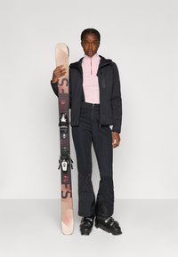 Czarna kurtka narciarska z zamkiem z przodu, różowa bluzka z długim rękawem, czarne spodnie narciarskie i czarne buty narciarskie, trzymająca brązową i różową nartę z wiązaniami.