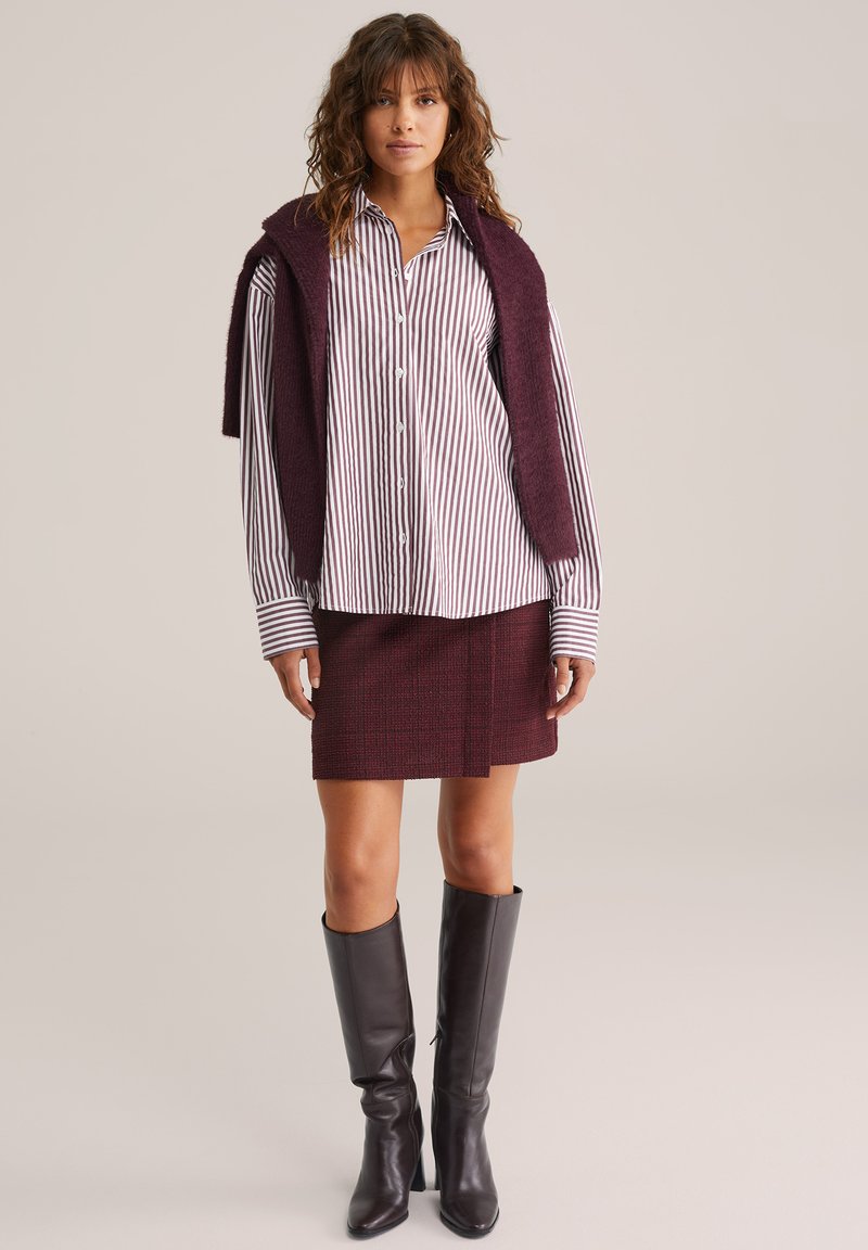 Camisa de botones a rayas en blanco y burdeos, combinada con una falda texturada burdeos a juego y botas de cuero negras hasta la rodilla.