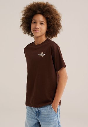 Jeune personne aux cheveux bouclés porte un t-shirt marron avec le texte « The Laidback Lounge » et un jean bleu clair, debout avec une main dans la poche.