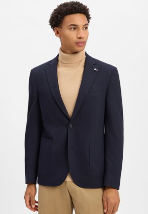 Man draagt een donkerblauw colbert over een beige coltrui en een kaki broek, staat tegen een effen witte achtergrond en kijkt naar rechts.