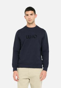 Marineblaues Sweatshirt mit Rundhalsausschnitt und gerippten Bündchen, das auf der Vorderseite ein auffälliges erhabenes Logo zeigt. Aus glattem Baumwollmischgewebe.