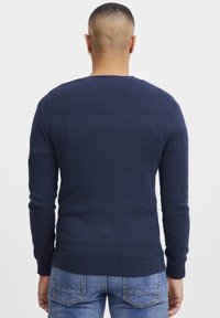 Mørkeblå strikkegenser med ribbestrikket skulderstruktur, tettsittende lange ermer og rund hals, sammen med lyseblå denimjeans.