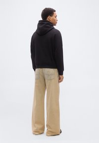 Άνδρας που κοιτάζει προς τα πίσω, φοράει μαύρο φούτερ με κουκούλα και μπεζ φαρδιά τζιν παντελόνια, στέκεται σε ένα απλό λευκό φόντο.