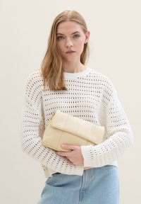 Borsa a mano in pelle crema con patta pieghevole; texture morbida e design minimalista. Modello che indossa un maglione bianco e jeans azzurri chiari.