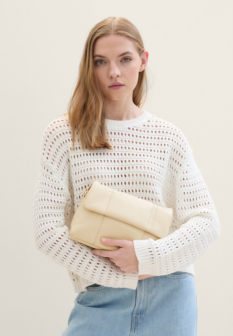 Borsa a mano in pelle crema con patta pieghevole; texture morbida e design minimalista. Modello che indossa un maglione bianco e jeans azzurri chiari.