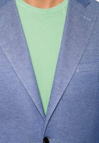 Svetle modrý blazer s texturovanou tkaninou a klopovými límci, v kombinaci s mátově zeleným tričkem pod ním, které má kulatý výstřih.