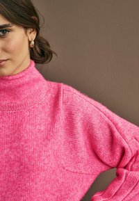 Maglione a collo alto rosa con vestibilità rilassata, lavorazione a maglia testurizzata e spalle scese. Presenta un aspetto morbido e accogliente su uno sfondo neutro.