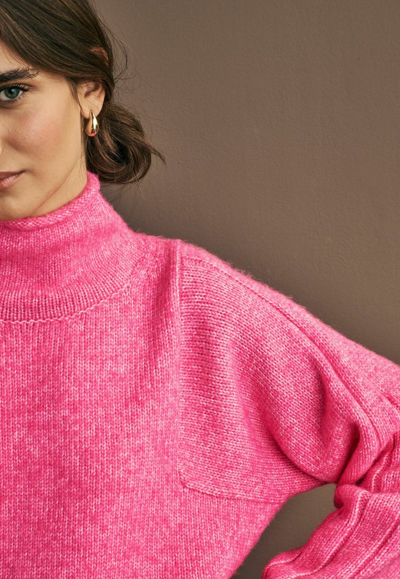 Maglione a collo alto rosa con vestibilità rilassata, lavorazione a maglia testurizzata e spalle scese. Presenta un aspetto morbido e accogliente su uno sfondo neutro.