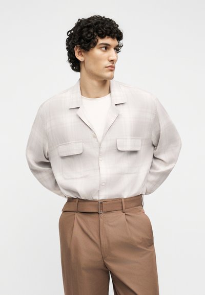 Άνδρας με σγουρά μαλλιά κοιτάζει δεξιά, φοράει ανοιχτό μπεζ πουκάμισο με κουμπιά πάνω από λευκό μπλουζάκι και καφέ παντελόνι ψηλόμεσο με ζώνη.