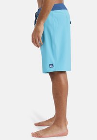 Shorts de bain bleus en matériau léger ; dotés d'une taille élastique avec un cordon de serrage bleu marine et un petit patch logo sur la jambe gauche.