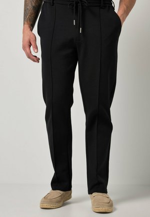 Mężczyzna noszący czarne spodnie zwężane z wiązanym w pasie ściągaczem oraz beżowe espadryle, stojący z rękami w kieszeniach.