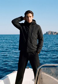 Homme portant une veste et un pantalon noirs se tient sur un bateau avec une main derrière la tête, océan et île rocheuse visibles en arrière-plan.