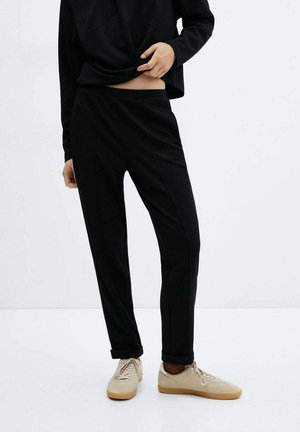 Personne portant un pantalon noir et un haut noir à manches longues, soulevant légèrement la chemise pour montrer la taille, avec des baskets beiges sur un sol blanc.