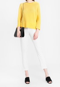 Geel gebreide trui, witte cropped broek met strepen, zwarte open tenen mules en een zwarte schoudertas met kettingriem.